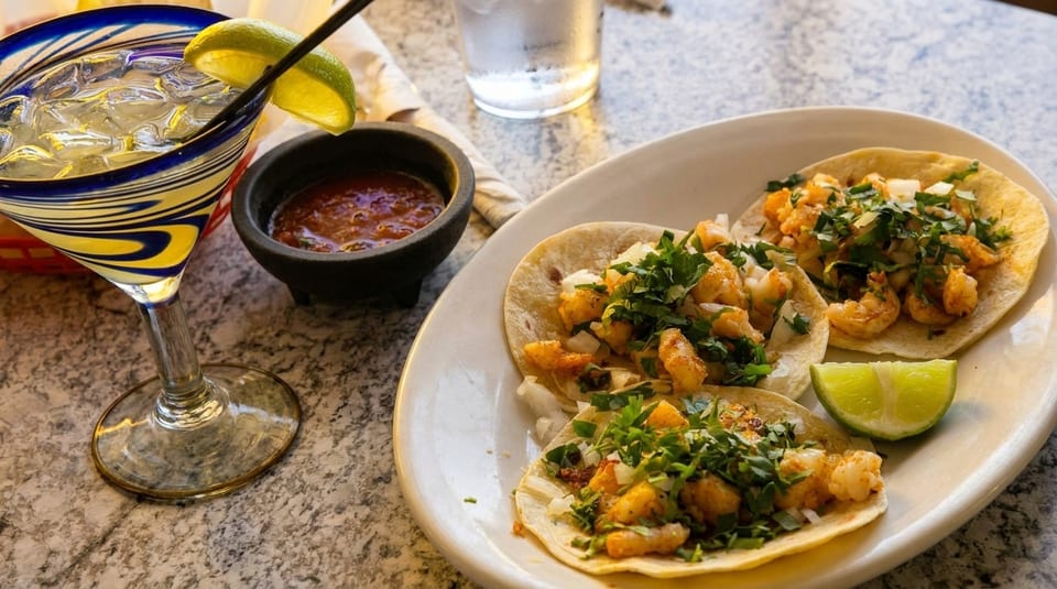 Shrimp tacos with cilantro, onions, and lime, alongside a margarita and salsa on a speckled table.
